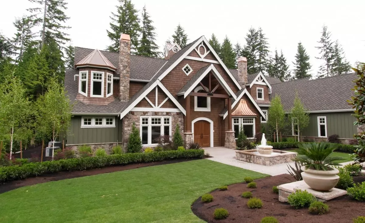 A Northwest craftsman custom home in Woodinville: cedar shingle gables with white timber trim, stone chimneys and a stone base, a copper-roofed turret and bay, an arched wood front door, and a fountain in a stone basin on the front walk.
