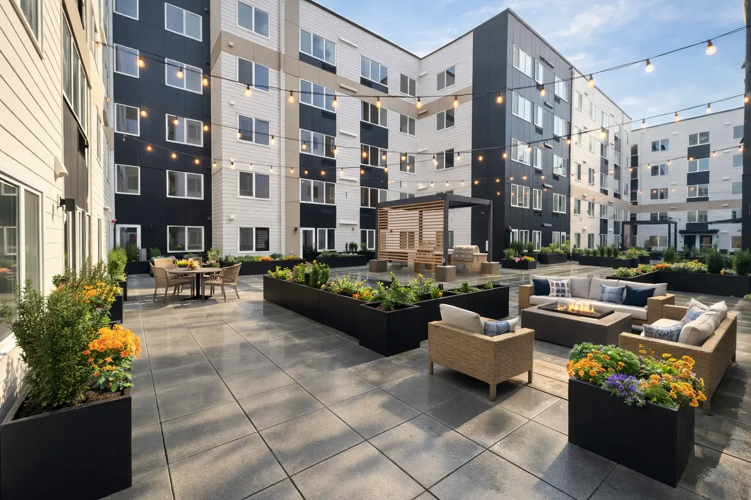 The courtyard at Enso Apartments in Lynnwood: five storeys of white lap siding with charcoal panel bays and taupe bands around a paved terrace with planters, a fire table, lounge seating and string lights.