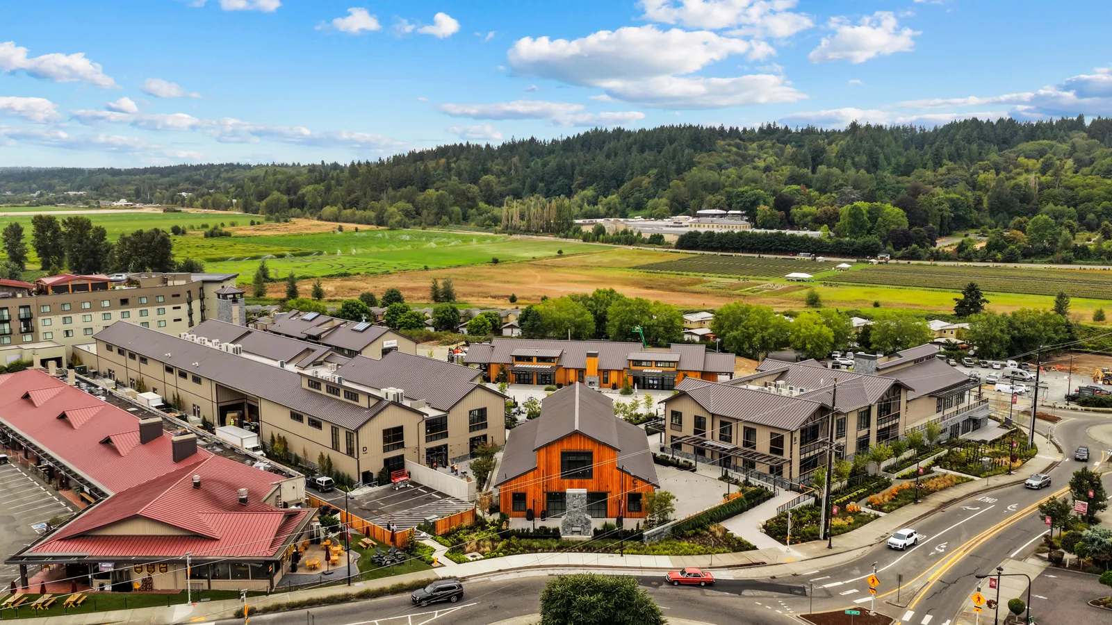 Harvest Wine Village in Woodinville from the air: a cluster of two-storey retail and tasting buildings with dark standing-seam metal roofs and pale board-and-batten siding, a barn-form building clad in vertical wood over a stone base at the centre, a red-roofed restaurant at the near corner, and newly striped parking and walkways between them, with farm fields, vineyard rows and forested hills behind.