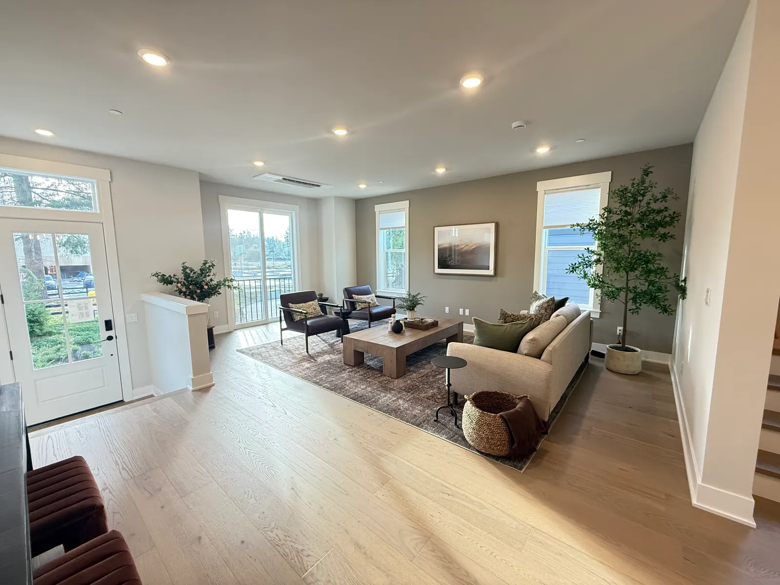 A bright open living room in warm white with light oak floors, a sofa and two leather chairs around a wood coffee table, a glass front door and tall windows on the left, and the stair rail on the right.