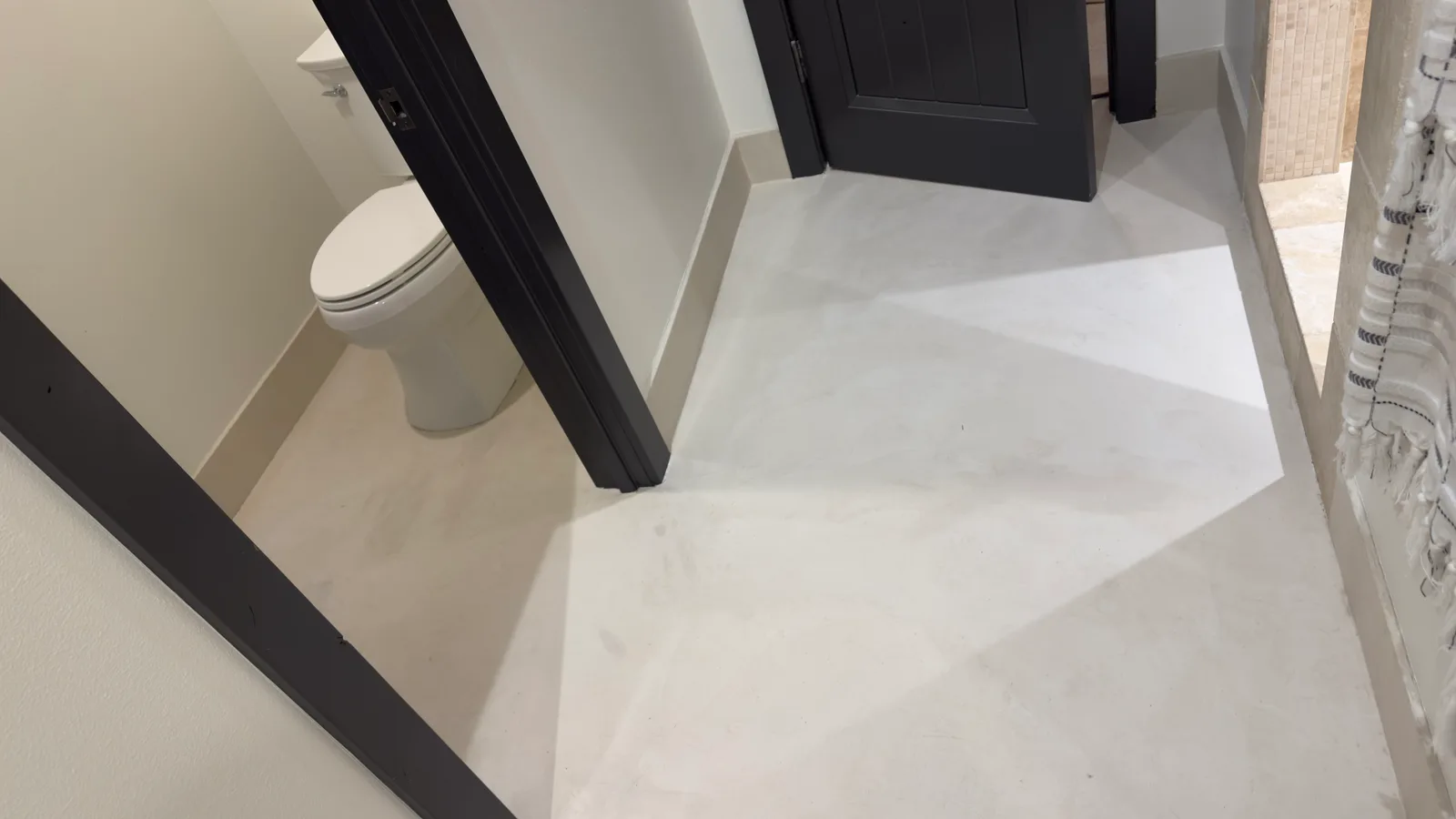 A finished microcement bathroom floor in pale warm grey, one seamless surface running from the vanity area through to the toilet room, framed by black door casings and cream baseboards.