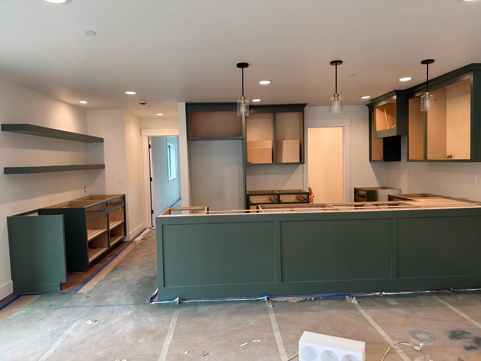 A Duvall kitchen mid-install with every cabinet finished in deep sage green: a long paneled island in front, upper cabinets on either side of a doorway, matching floating shelves and a base cabinet run on the left wall, and glass pendant lights over the island.