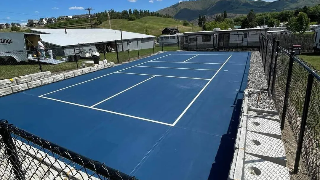A new pickleball court coated in deep blue with crisp white lines, set behind black chain-link fencing and concrete block walls, with green hills and a blue sky beyond.