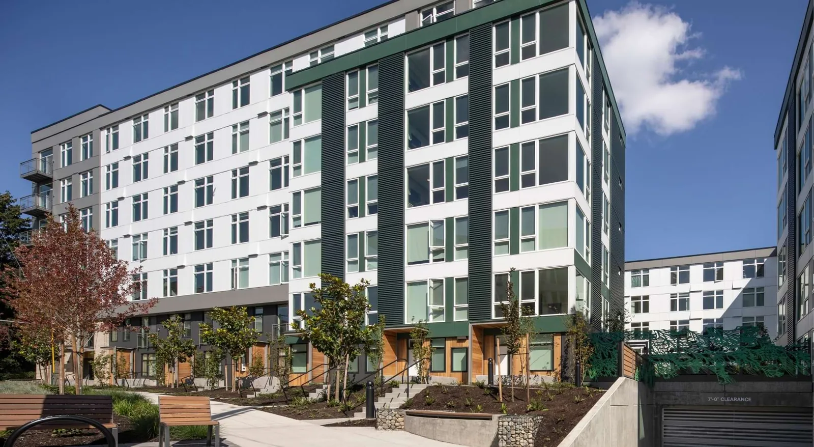 Wayfarer Apartments in Seattle: a seven-storey building in white panels with dark green corrugated bays and window frames, cedar-clad ground-floor entries, and a newly planted courtyard with benches in front.
