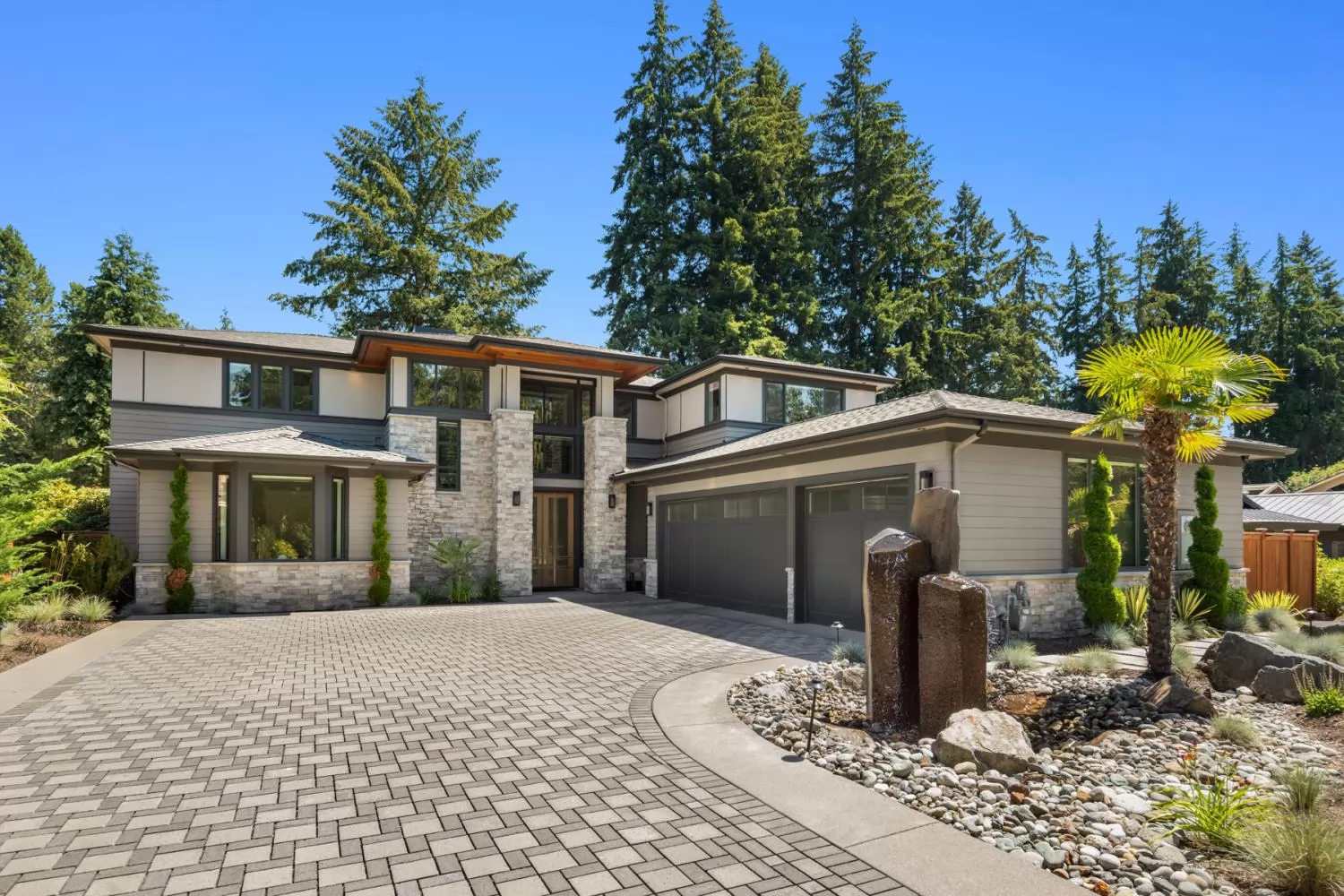A two-storey Northwest contemporary custom home by Stackman Custom Homes: grey lap siding with dark trim, a stacked-stone entry tower, cedar soffits under low hipped roofs, a two-car garage in charcoal, and a paver drive curving past a rock garden and palm under tall firs.