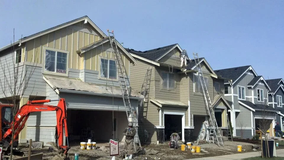 A row of new production homes mid-paint: RC painters on extension ladders at the rooflines and another at an open garage, paint buckets and a compact excavator on the bare ground in front, and SOLD signs in the upper windows.